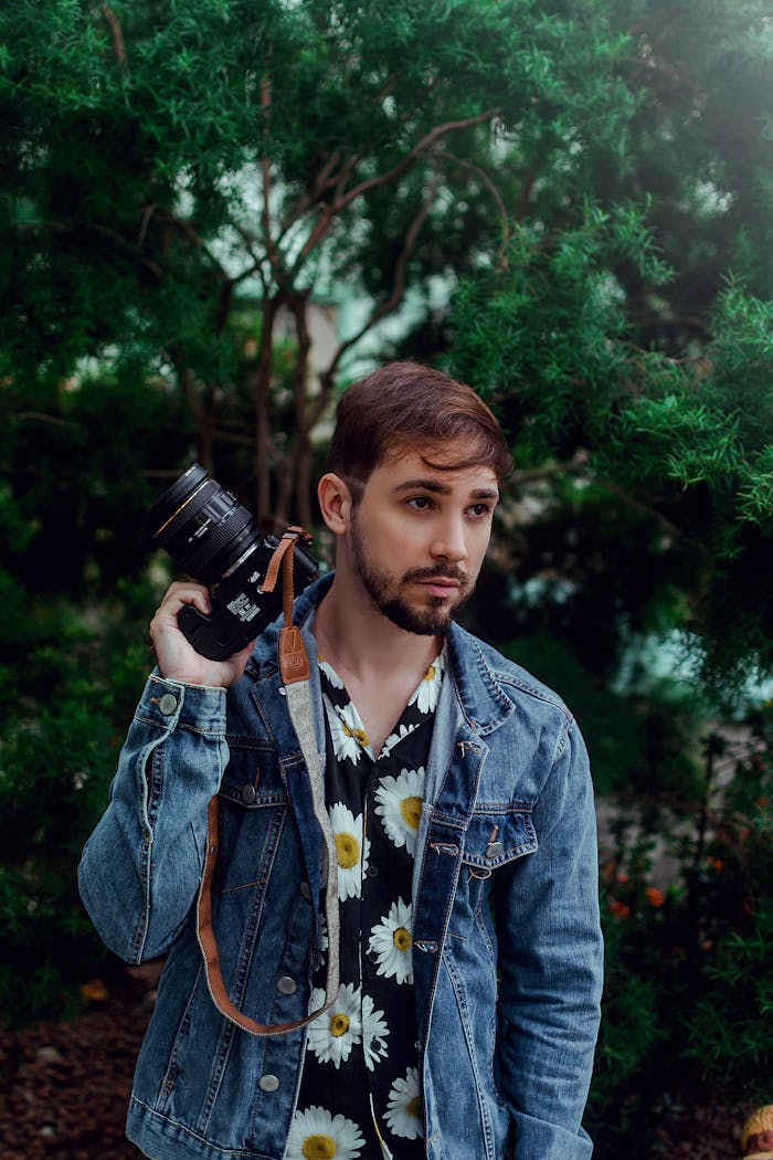 A fashionable man in a floral shirt and denim jacket poses with a camera against a lush green backdrop.