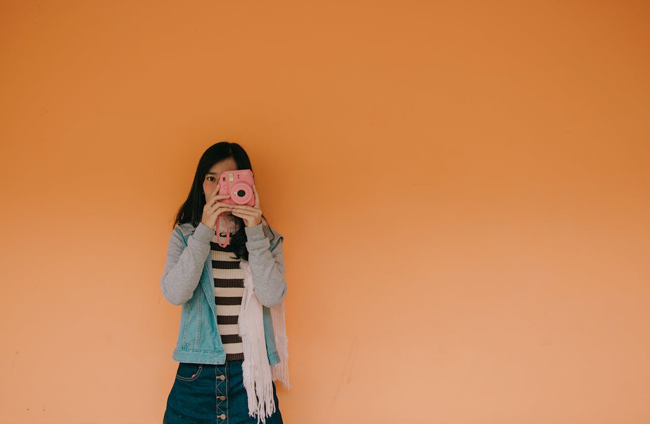 Young woman holding a pink camera stands against an orange wall, exuding a playful fashion vibe.